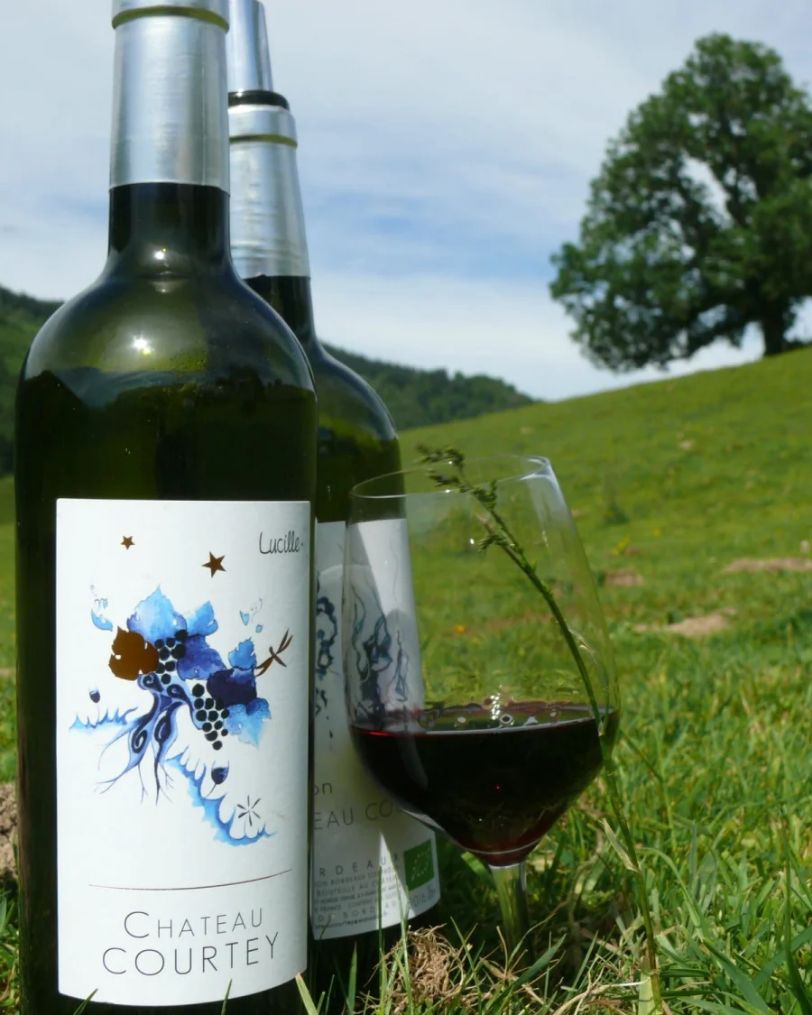 Deux bouteilles de Château Courtey, dont la cuvée Lucille, et un verre de vin rouge posés dans l'herbe devant un pré et un grand arbre